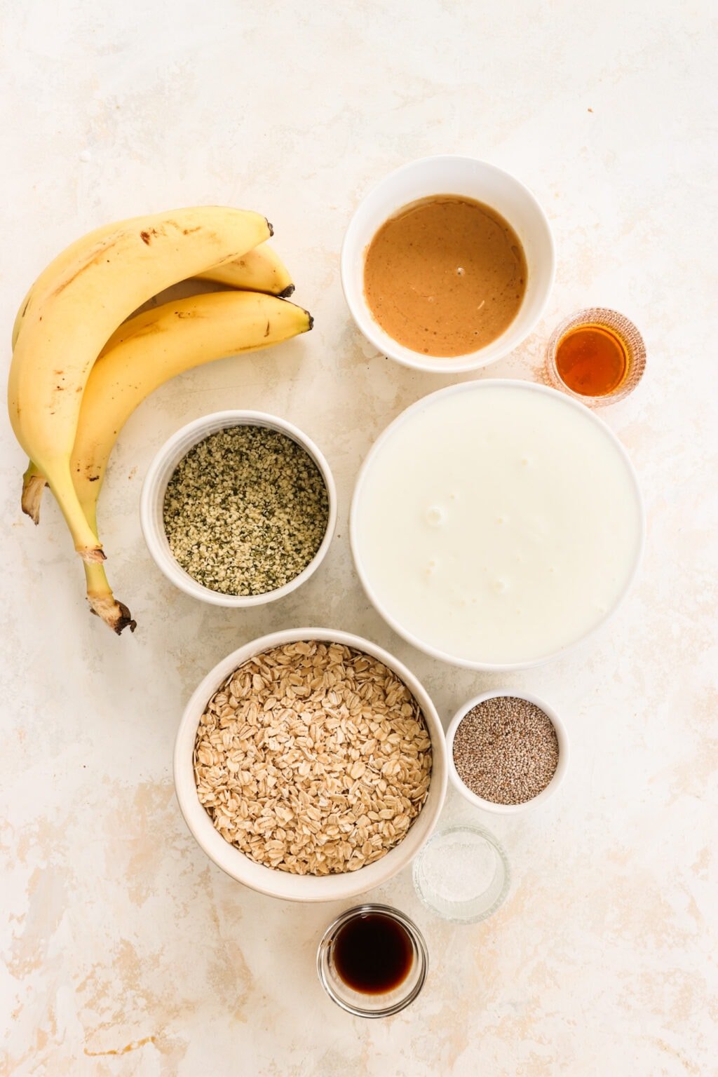 Ingredients for Peanut Butter & Banana Kefir Overnight Oats in glass bowls, including quick oats, hemp hearts, chia seeds, vanilla extract, plain kefir, maple syrup, peanut butter, and bananas