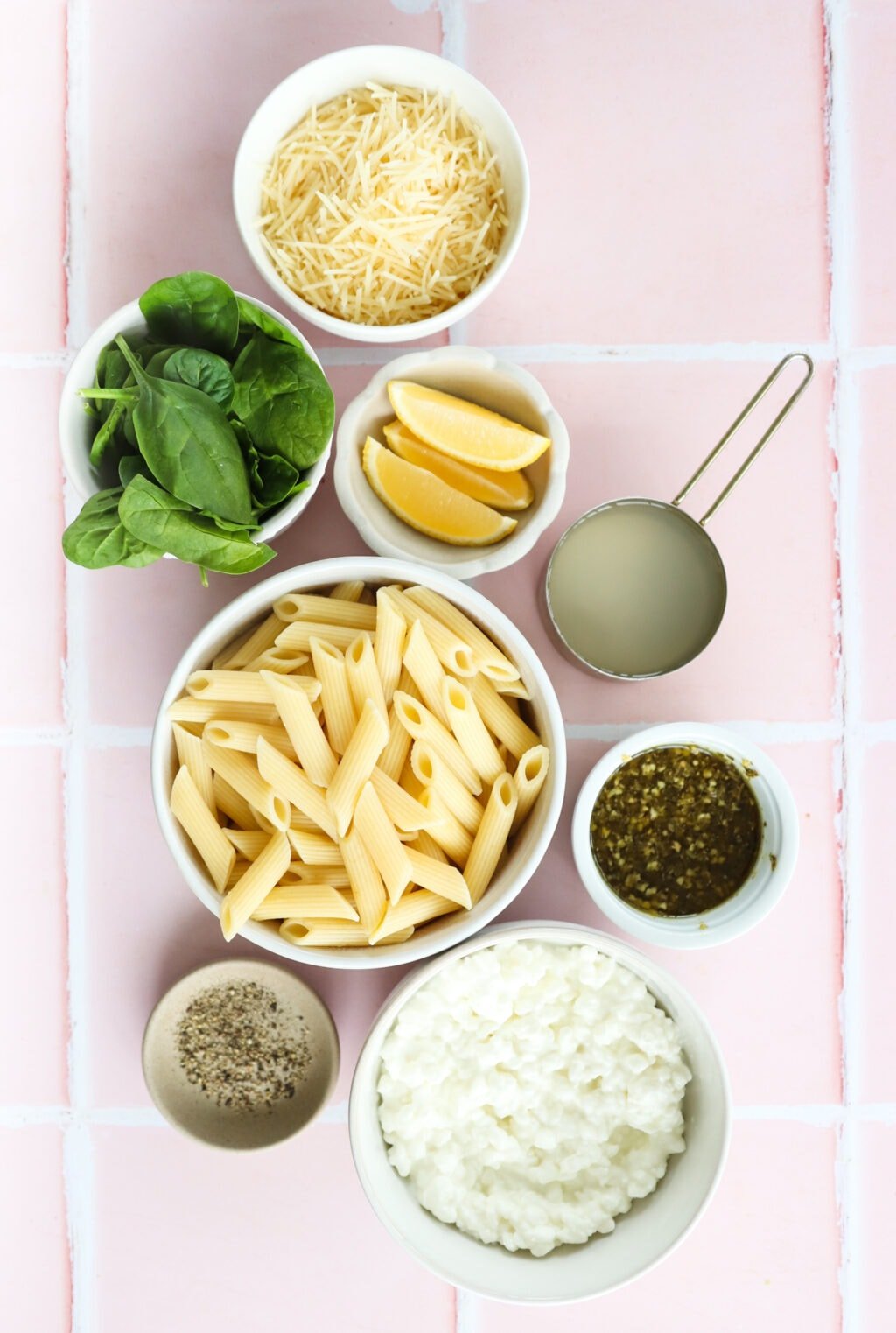 Ingredients for 20 minute pesto cottage cheese pasta in small white bowls, including pasta noodles, pasta water, cottage cheese, pesto, spinach, salt and pepper, parmesan cheese, lemon wedges