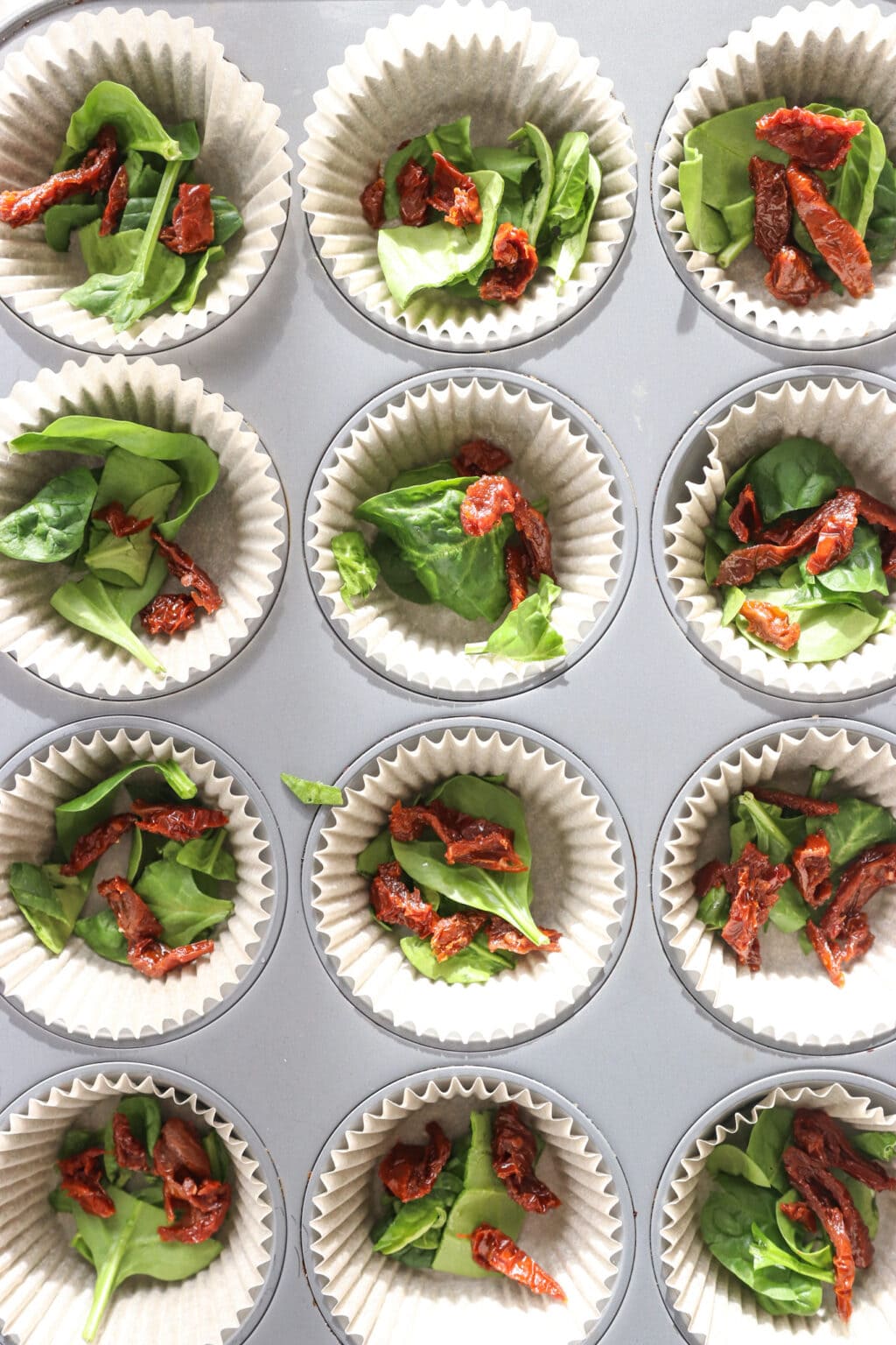 Ingredients being prepared in a muffin tin for Egg Bites with Cottage Cheese, Spinach & Sun-dried Tomatoes, including the sun dried tomatoes and spinach
