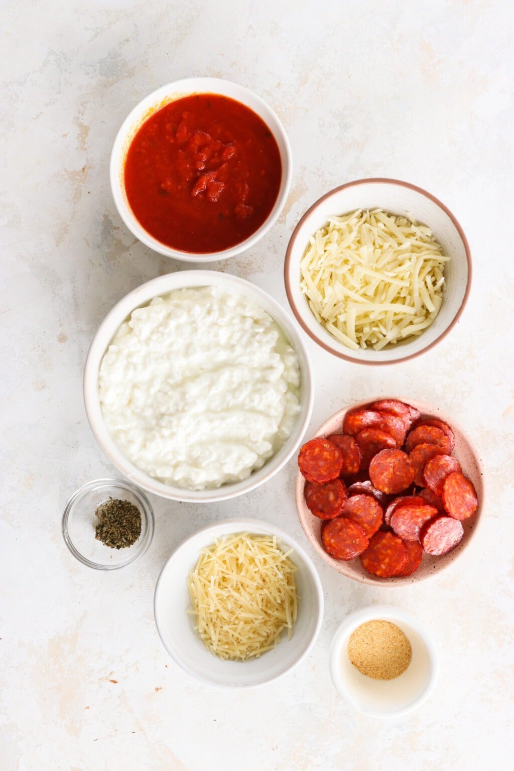 Ingredients for Quick Baked Cottage Cheese Pizza Bowl Dip in white bowls, including cottage cheese, parmesan cheese, garlic powder, Italian seasoning, marinara sauce, mozzarella cheese, turkey pepperoni or pepperoni