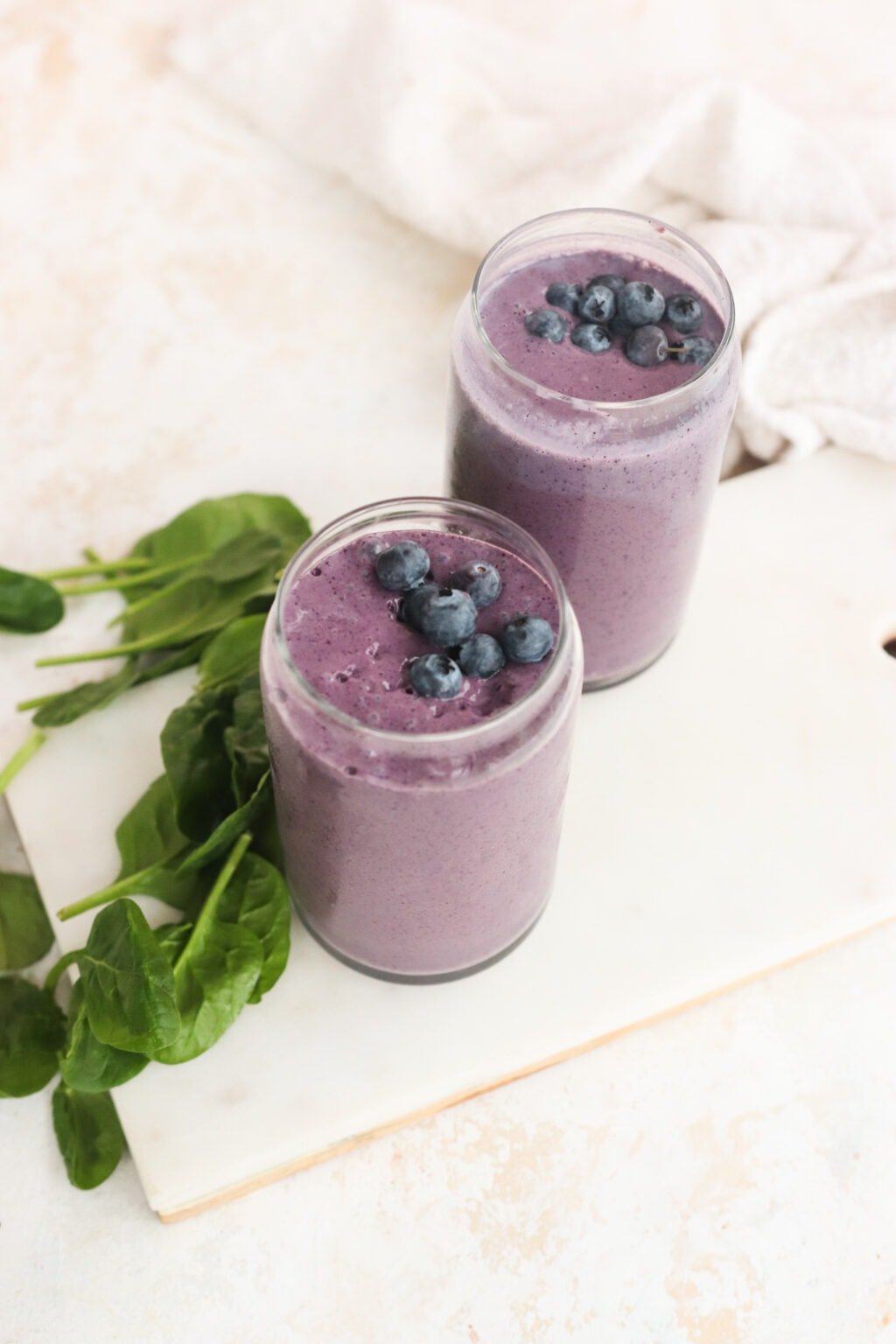 Two clear glasses filled with High Protein Blueberry Breakfast Smoothie with Cottage Cheese on a cutting board with spinach on the side