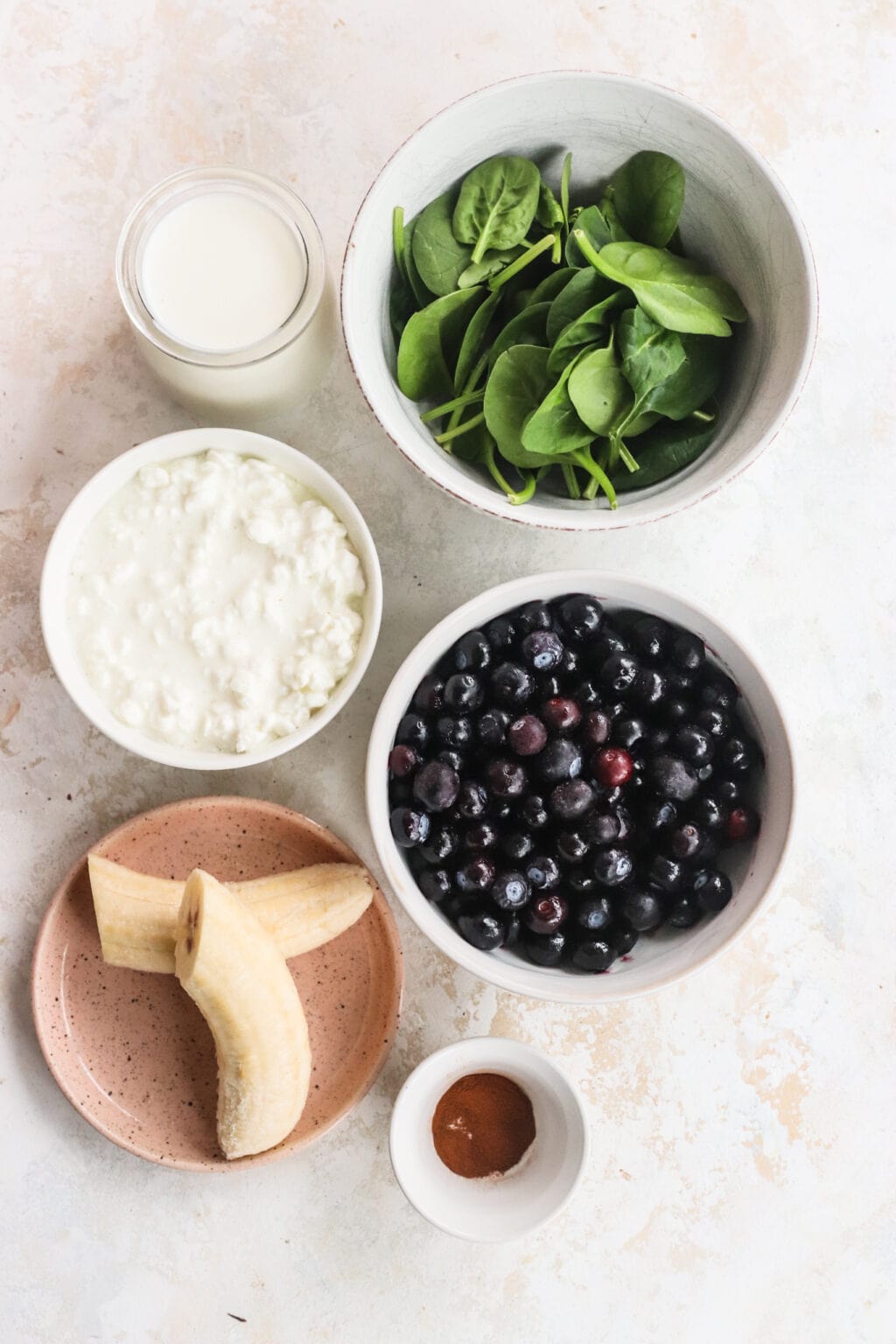 Ingredients for High Protein Blueberry Breakfast Smoothie in small bowls, including blueberries, banana, cottage cheese, milk, cinnamon, spinach, chia seeds, hemp hearts