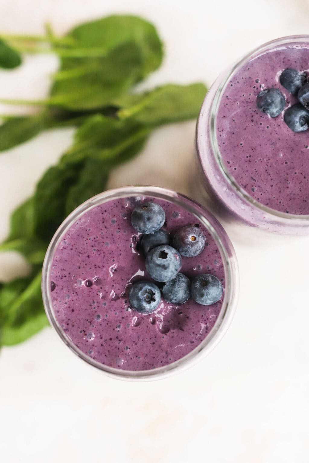 Two clear glasses filled with High Protein Blueberry Breakfast Smoothie with Cottage Cheese on a cutting board with spinach on the side