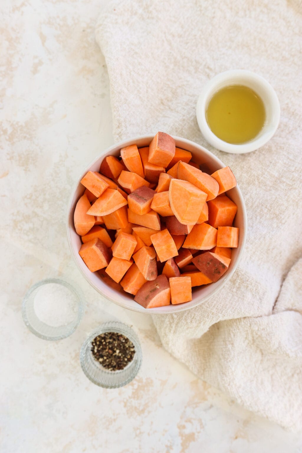 Ingredients for roasted sweet potatoes in white and clear glass bowls, including sweet potatoes, avocado oil, salt, freshly ground black pepper