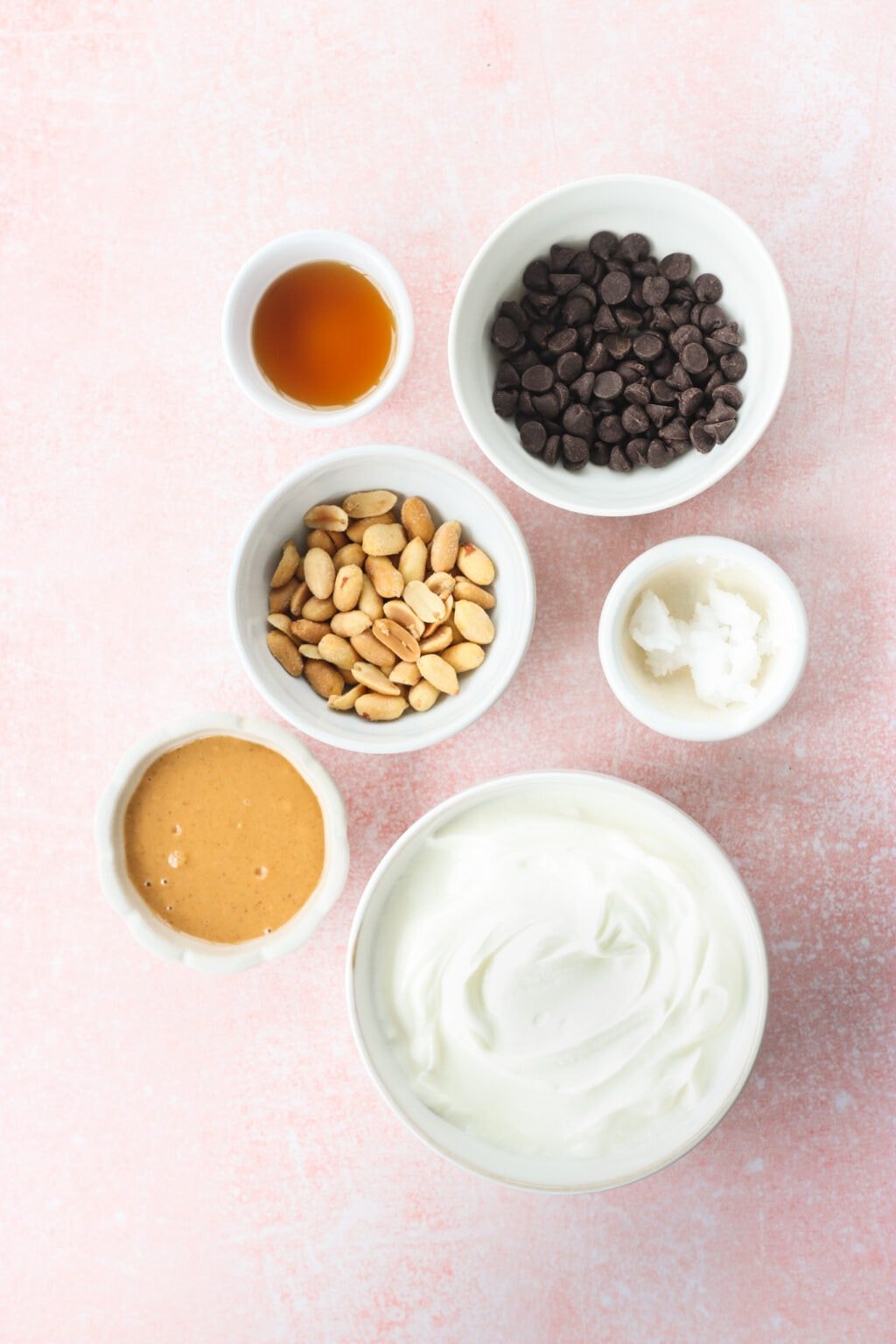 Ingredients for Peanut Butter & Greek Yogurt Cups with Magic Chocolate Shell in small white bowls on a pink marble counter, including Greek yogurt, smooth peanut butter, maple syrup, salted toasted peanuts, chocolate, coconut oil