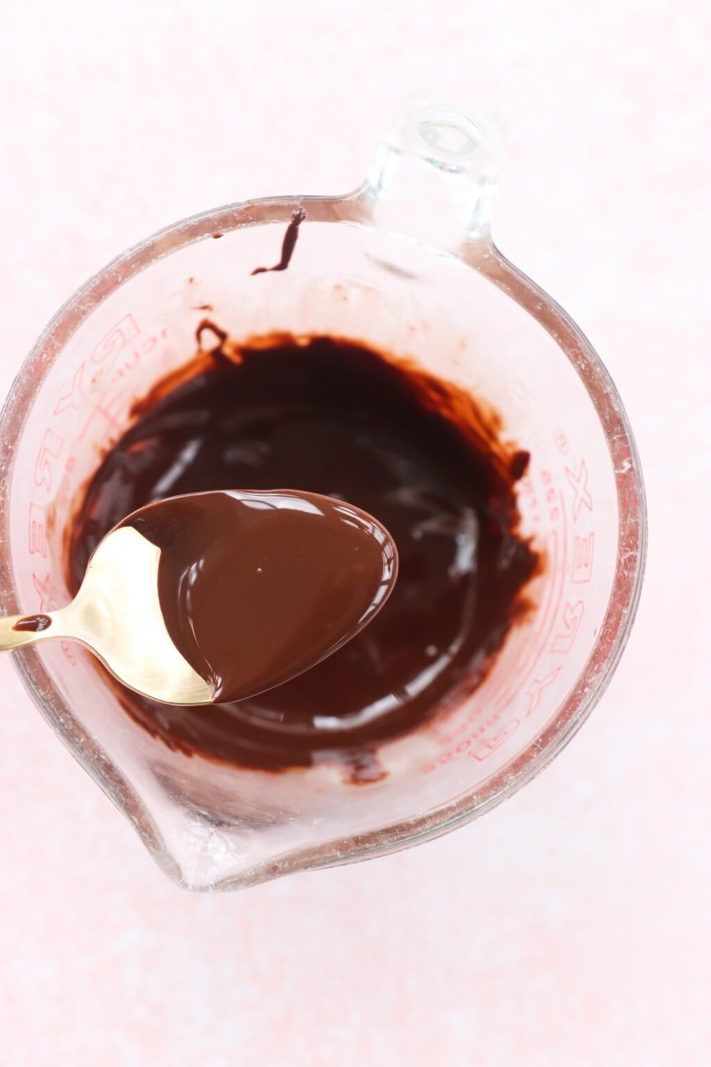 Ingredients for Peanut Butter & Greek Yogurt Cups with Magic Chocolate Shell mixed in a glass measuring cup on a pink marble counter, including chocolate and coconut oil