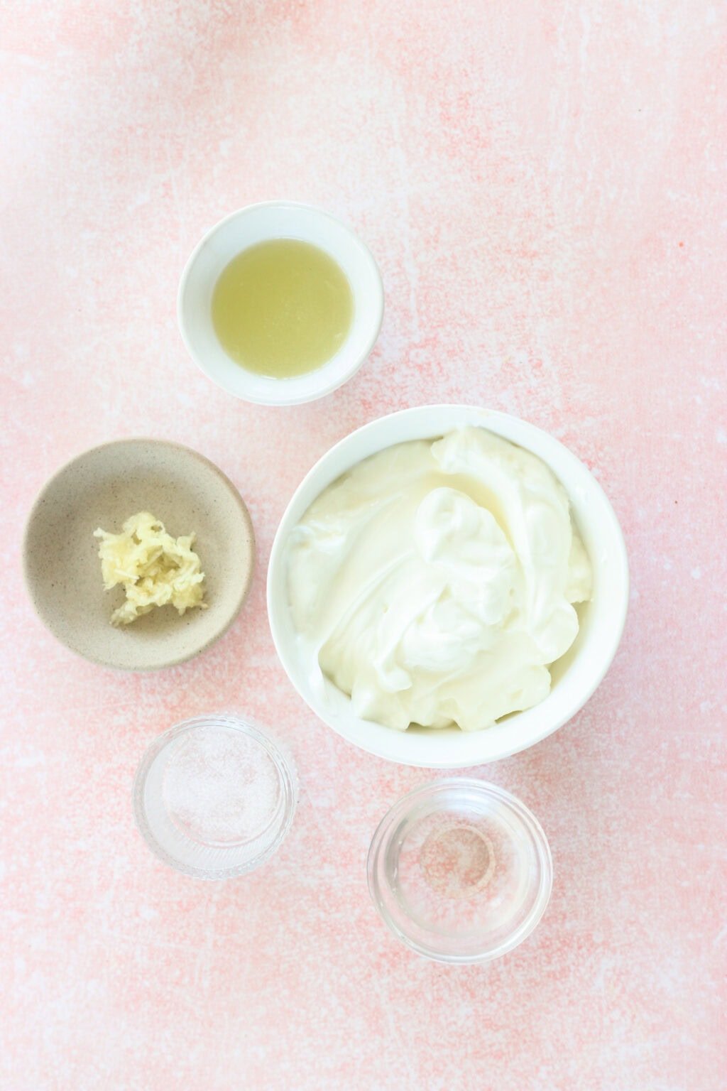 Ingredients for 4 Ingredient Greek Yogurt Lime Crema in small white bowls on a pink countertop, including lime juice, Greek yogurt, garlic, salt, and warm water