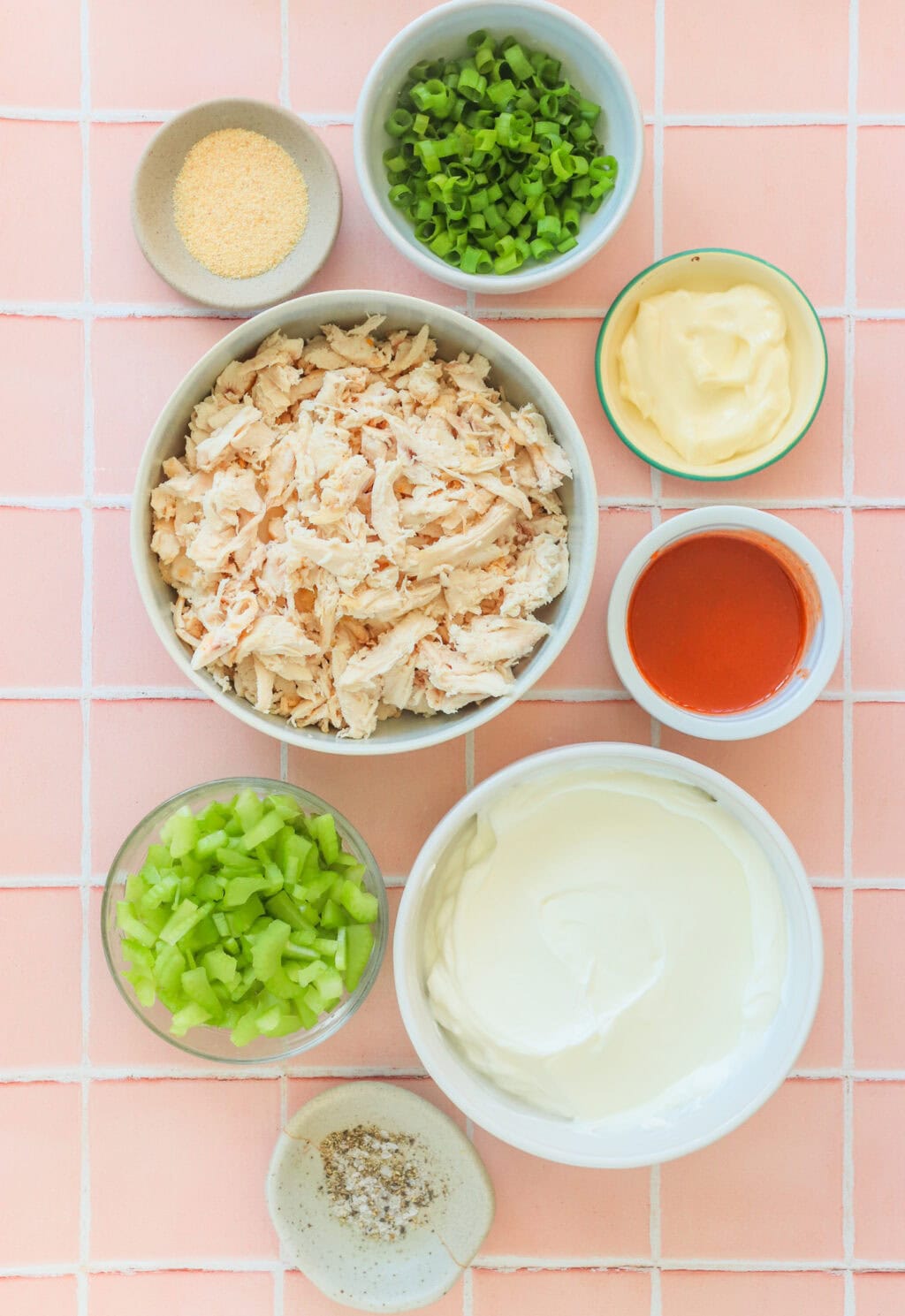 Ingredients for Quick Buffalo Chicken Salad with Greek Yogurt in small white bowls including cooked shredded chicken, Greek yogurt, mayo, Frank's Red Hot Original Sauce, garlic powder, green onions, blue cheese, pepper