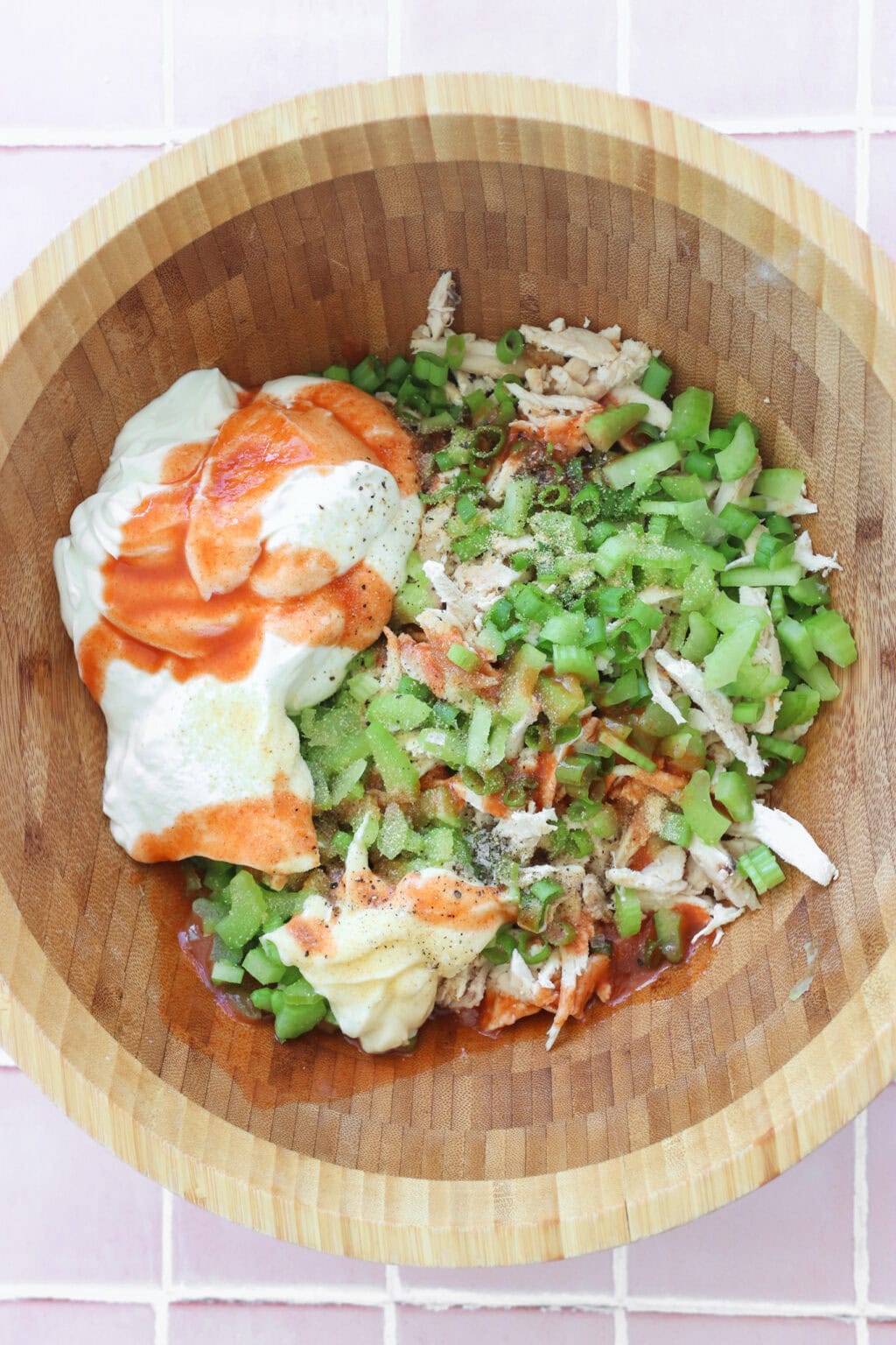 Ingredients for Quick Buffalo Chicken Salad with Greek Yogurt unmixed in a medium wooden bowl including cooked shredded chicken, Greek yogurt, mayo, Frank's Red Hot Original Sauce, garlic powder, green onions, blue cheese, pepper