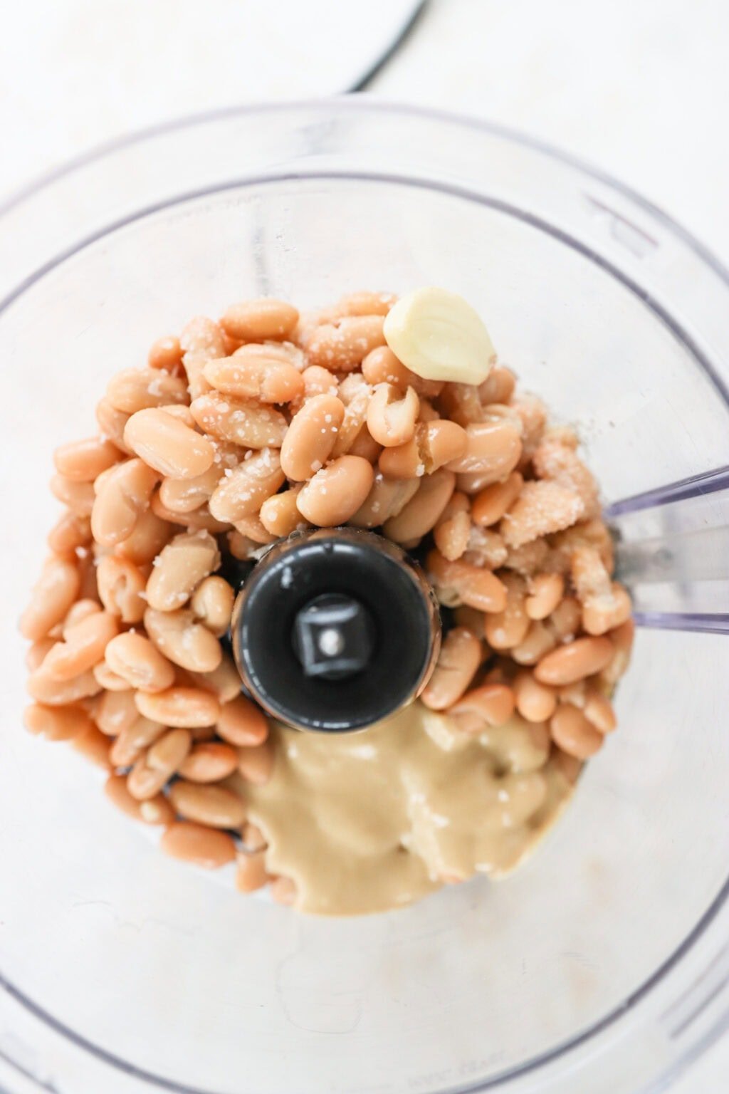 Ingredients for Super Creamy Vegan White Bean Dip with Cashews unblended in a food processor, including cannellini beans, cashews, extra virgin olive oil, salt, lemon juice