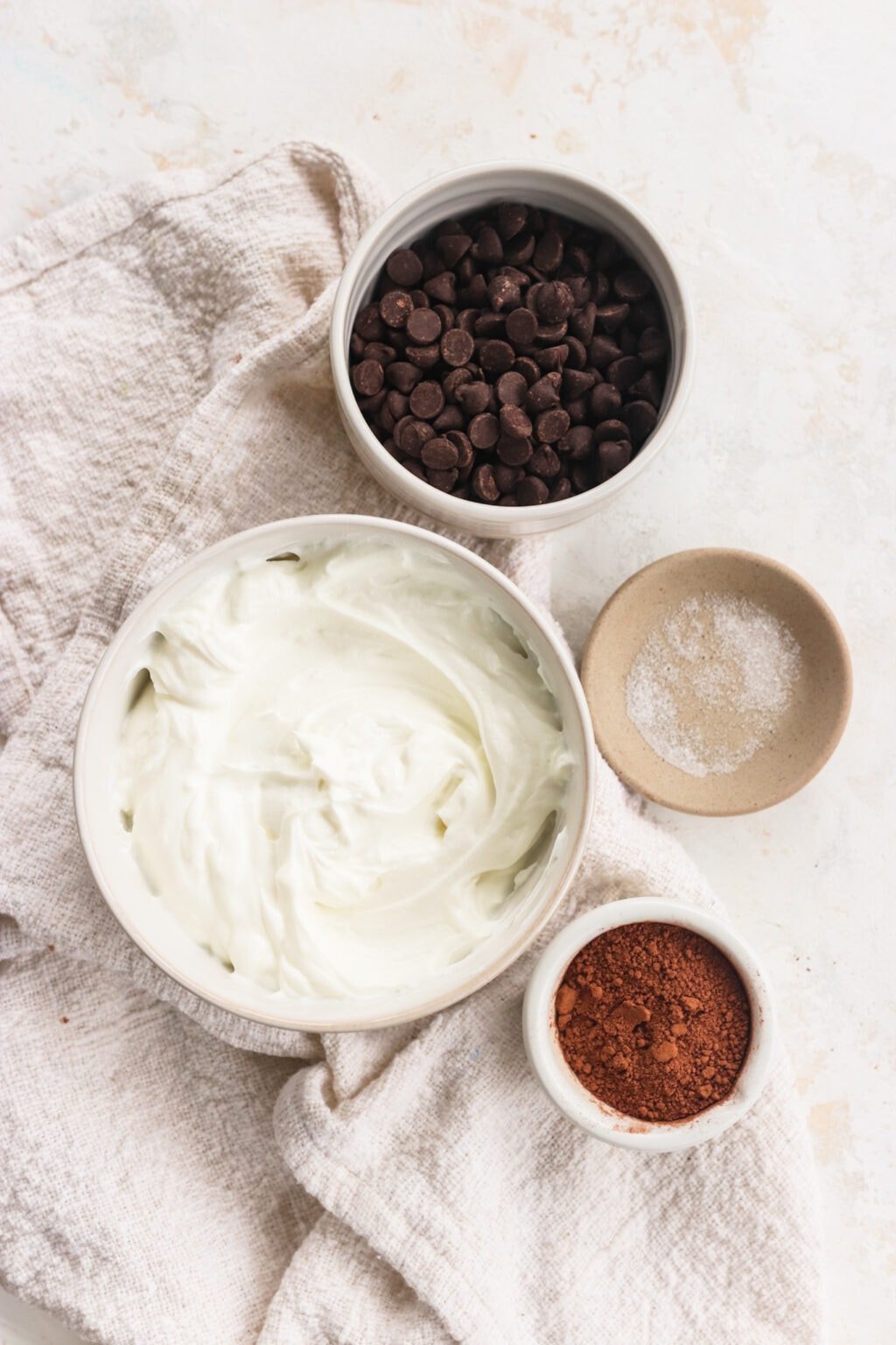 Ingredients for Whipped Greek Yogurt Chocolate Mousse in white glass bowls, including chocolate chips, Greek yogurt, cacao and salt