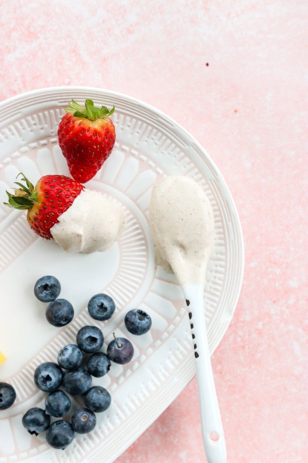A white plate on a pink countertop with blueberries, strawberries, and a spoon that has been dipped in The Best Honey-Cinnamon Greek Yogurt Fruit Dip (easy high protein snack)