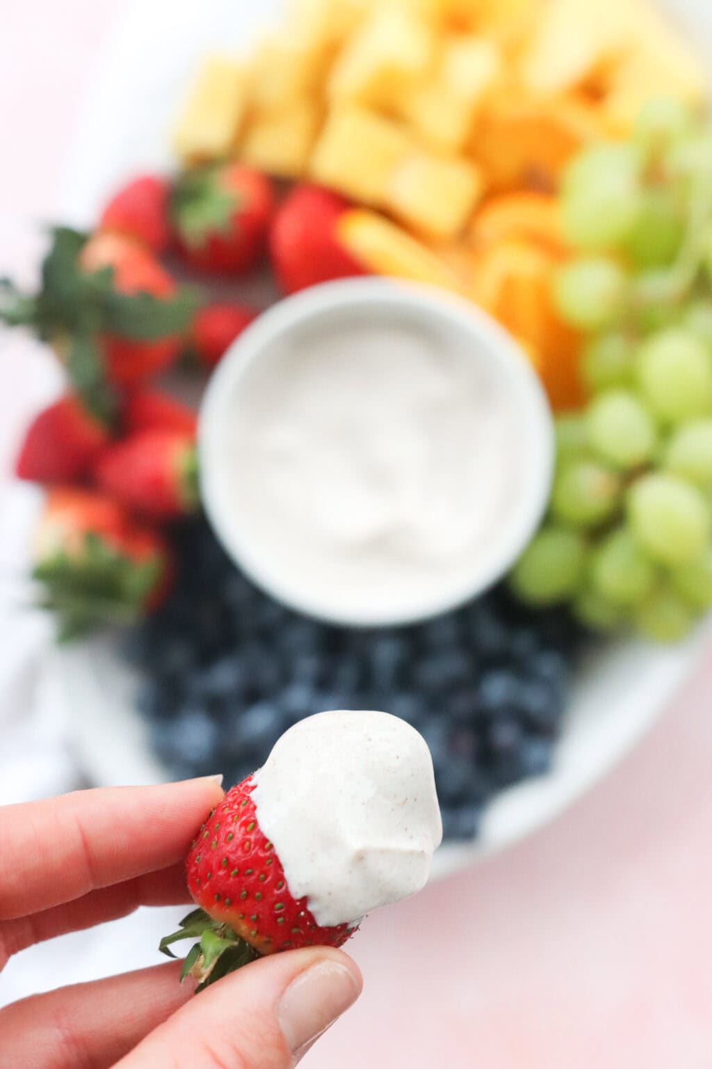 A close up of a strawberry dipped in the Best Honey-Cinnamon Greek Yogurt Fruit Dip (easy high protein snack) in a white bowl on top of a tray filled with fruit