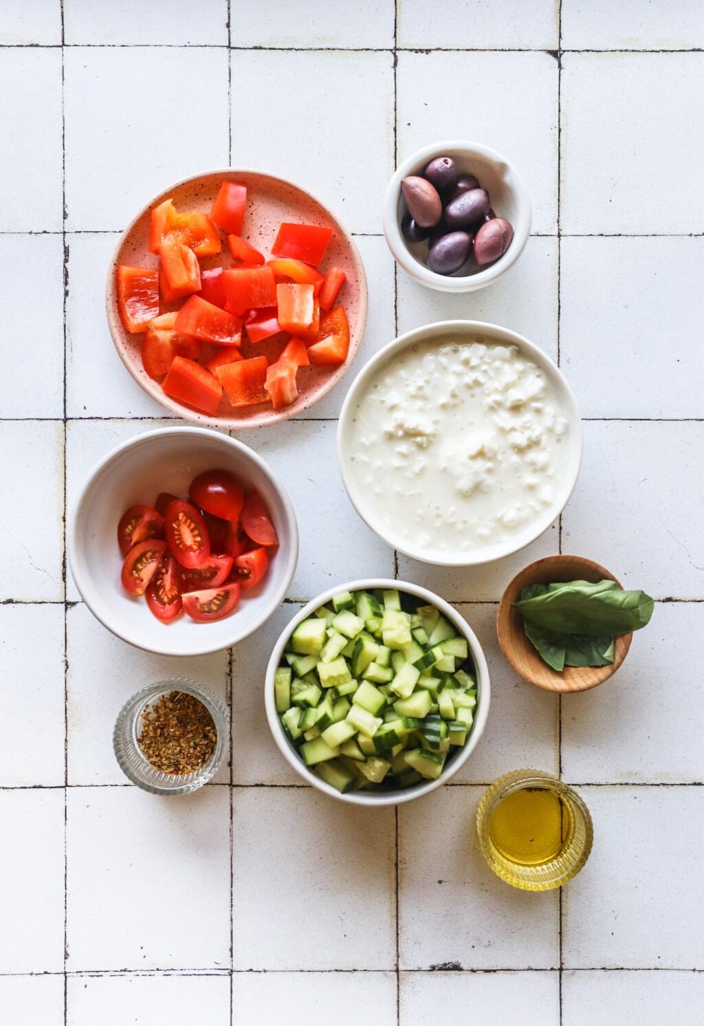 Ingredients for Easy Greek-Inspired Cottage Cheese Salad in small white bowls, including cottage cheese, cherry tomatoes, cucumber, red peppers, olives, Greek seasoning, olive oil, fresh dill
