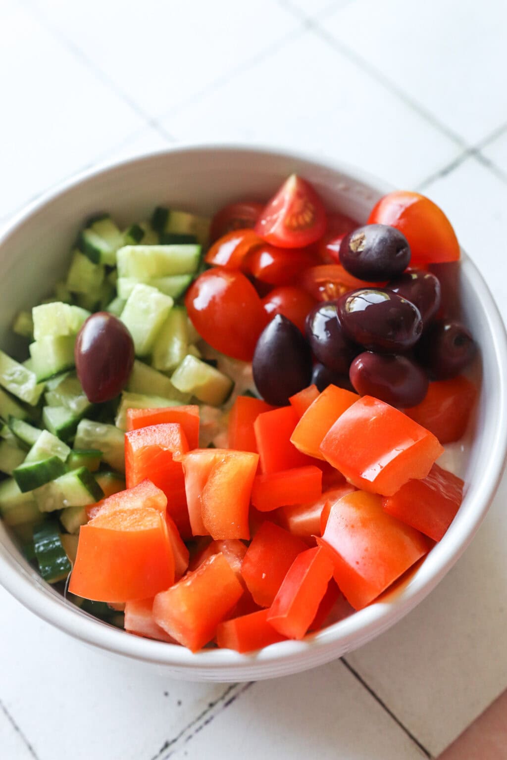 Ingredients for Easy Greek-Inspired Cottage Cheese Salad unmixed in a white bowl, including cherry tomatoes, cucumber, red peppers, olives