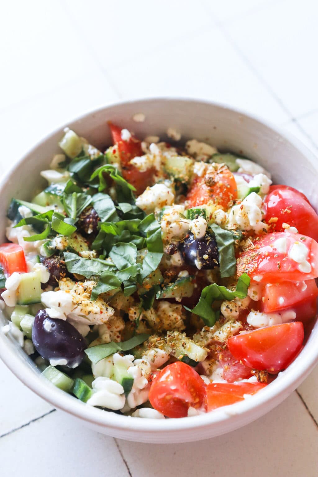 Ingredients for Easy Greek-Inspired Cottage Cheese Salad mixed in a white bowl, including cherry tomatoes, cucumber, red peppers, olives