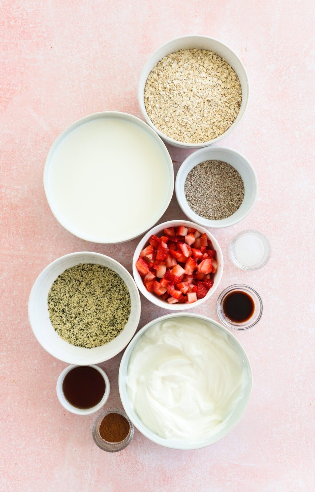 Ingredients for Grab-and-Go Strawberry Overnight Oats in small white bowls, including rolled oats, milk, maple syrup, chia seeds, hemp hearts, ground cinnamon, salt, strawberries, vanilla Greek yogurt