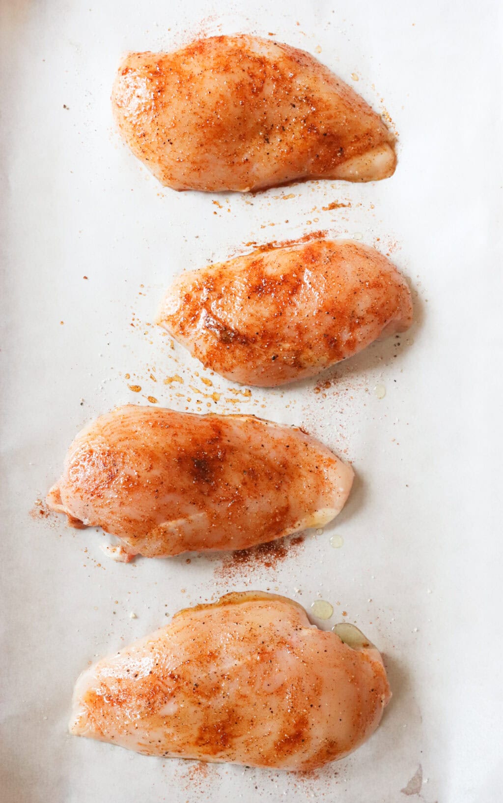 Four uncooked seasoned chicken breasts on a baking sheet with parchment paper