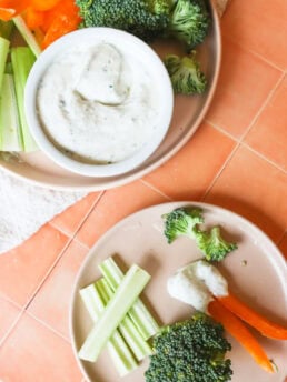 final image of the high protein cottage cheese ranch dip, with celery, peppers and broccoli on a light pink plate, on an orange countertop, and a smaller plate with a few celery sticks, broccoli and peppers dipped in the dip