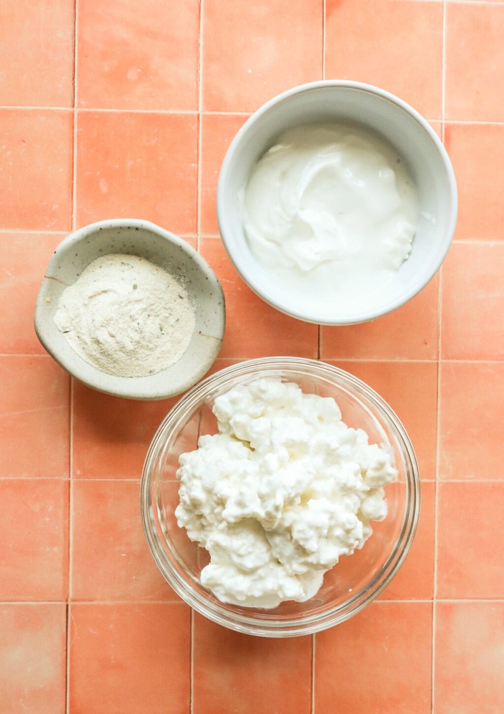 Ingredients to make high protein cottage cheese ranch dip in three separate bowls on a orange countertop, glass bowl with cottage cheese, white bowl with greek yogurt and small bowl with ranch seasoning
