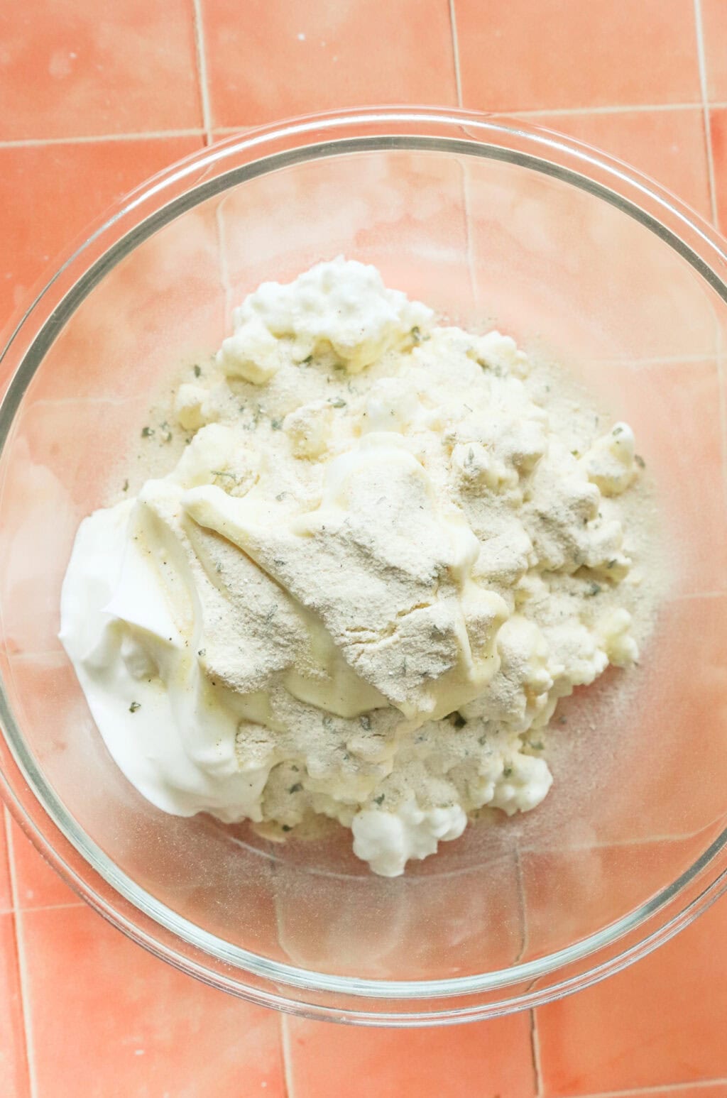 ingredients in a glass bowl, on orange countertop, ingredients include; cottage cheese, ranch seasoning pack, salt, and greek yogurt