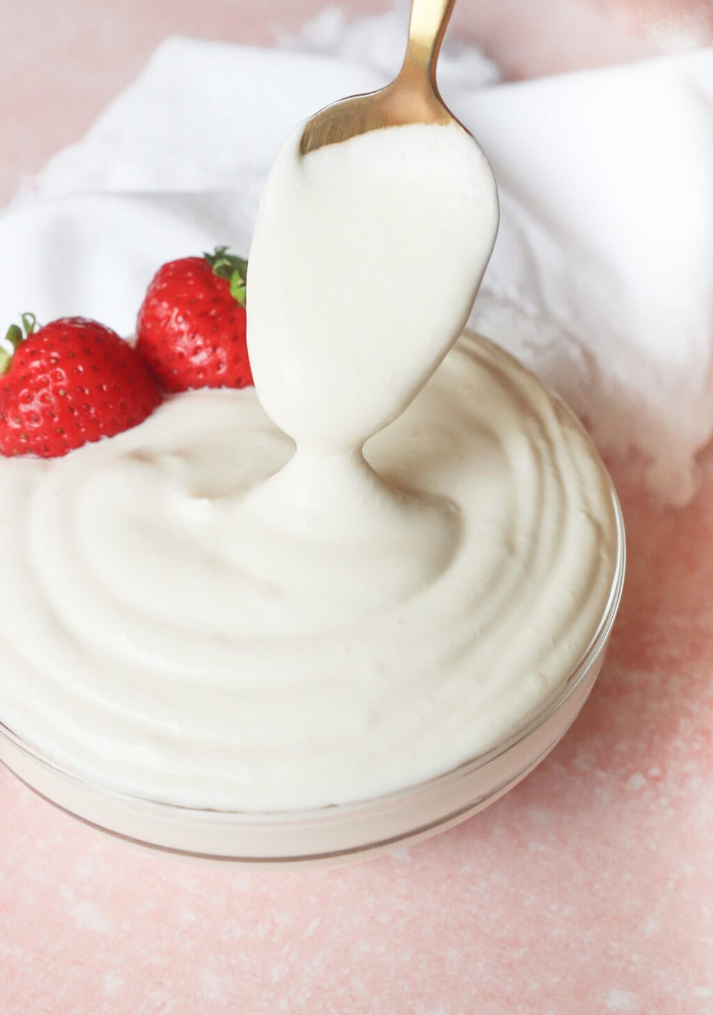 A spoon scooping a generous amount of creamy, thick Greek yogurt whipped cream from the bowl, with fresh strawberries on the side for garnish.