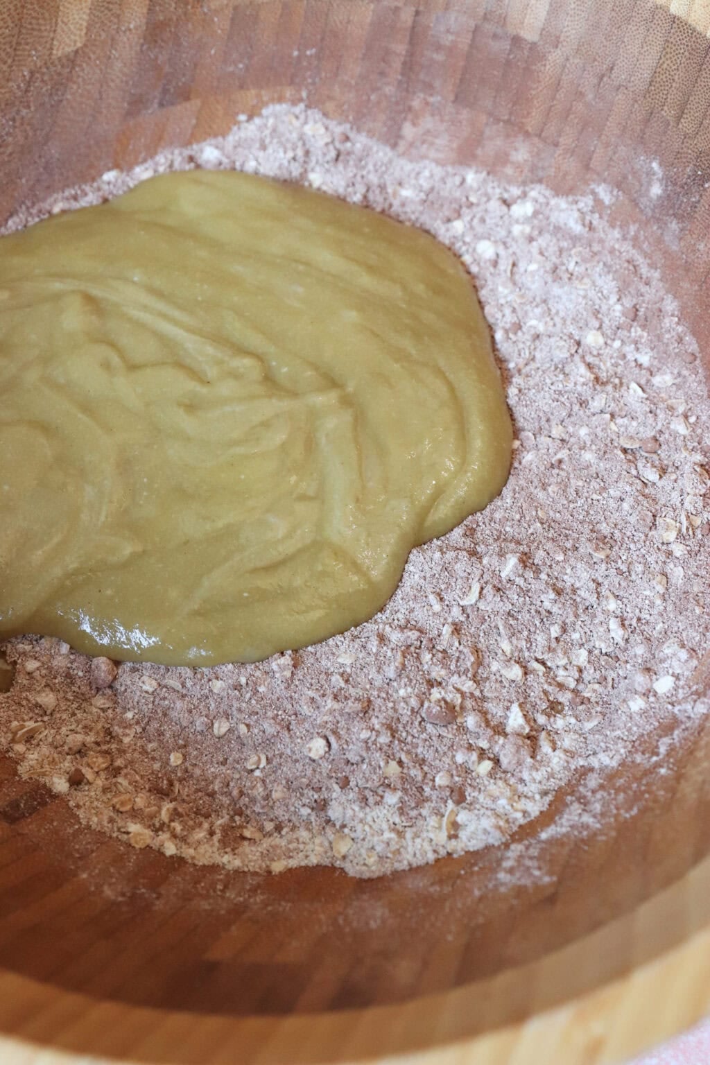 Batter is being mixed in a wooden bowl, with a wet mixture poured over a heap of dry ingredients, likely for baking healthy breakfast cookies.