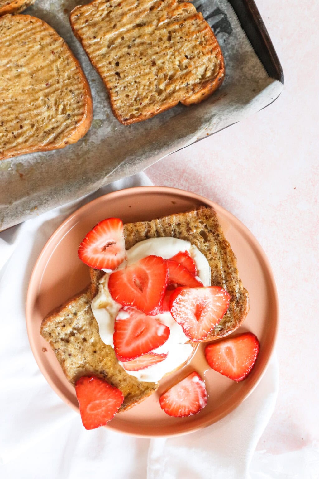 final product of sheet pan french toast with whipped greek yogurt and strawberries on top on a pink plate with some other slices on the french toast in the background