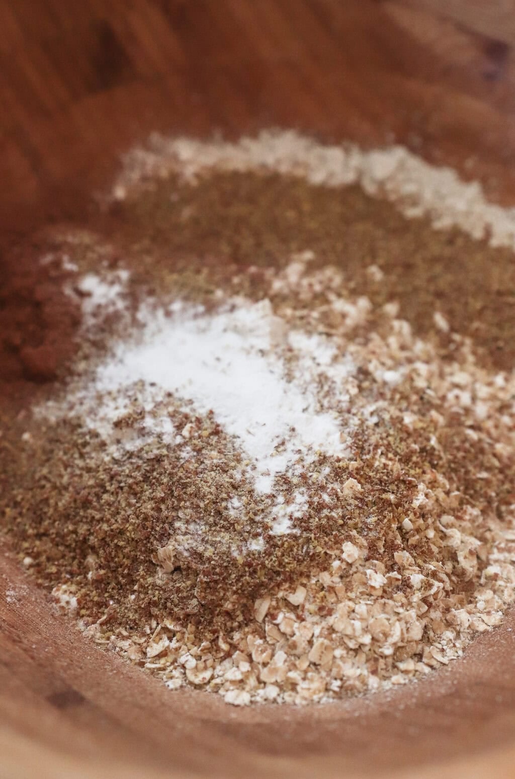 A close-up of a wooden bowl containing a mix of dry baking ingredients—oats, ground flaxseed, flour, baking powder, and cinnamon—ready to be combined for healthy breakfast cookies.