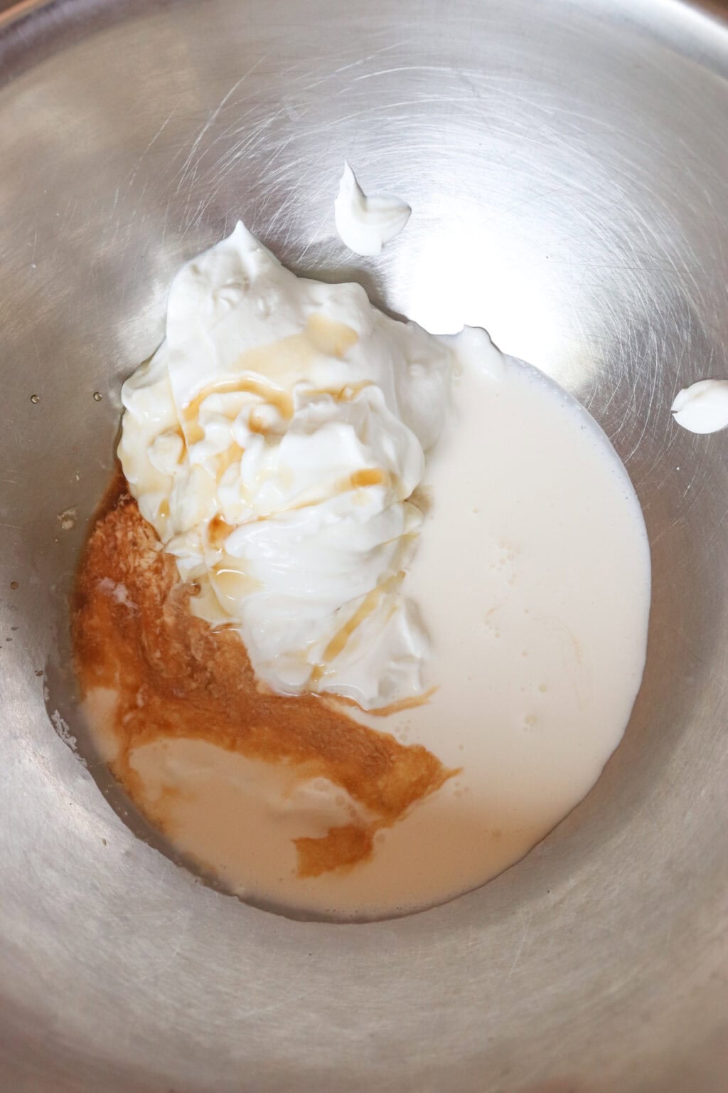 A stainless steel mixing bowl containing a blend of Greek yogurt, cream, vanilla extract, and sweetener, ready for whipping