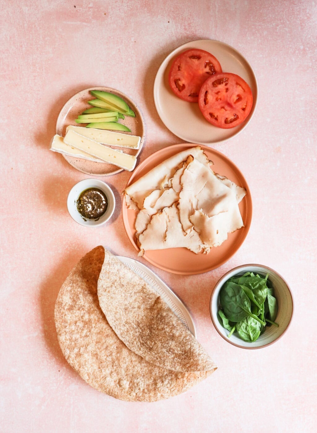all the ingredients for the turkey pesto wrap, spinach, whole wheat wrap, turkey, pesto, cheese, tomato, and avocado