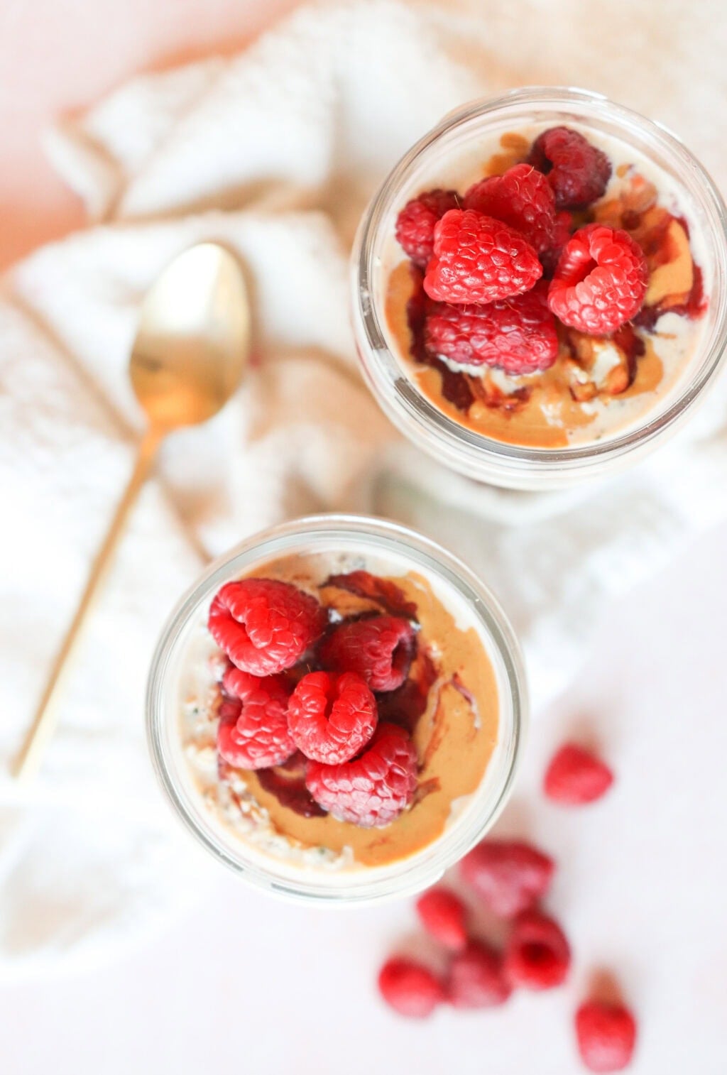 A top-down view of two jars of peanut butter and jelly overnight oats, both topped with fresh raspberries and peanut butter drizzle, with a golden spoon and scattered raspberries in the background.