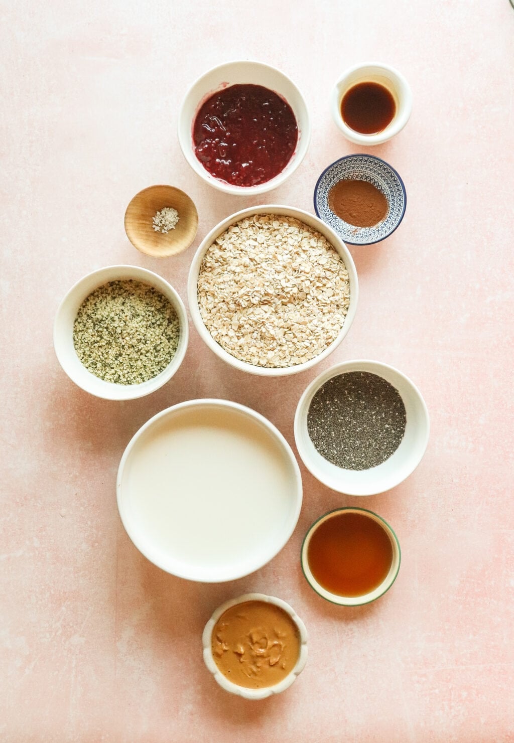 An assortment of ingredients for making PB&J overnight oats, including oats, peanut butter, chia seeds, raspberry jam, maple syrup, cinnamon, and almond milk, neatly arranged in small bowls on a pastel pink surface.