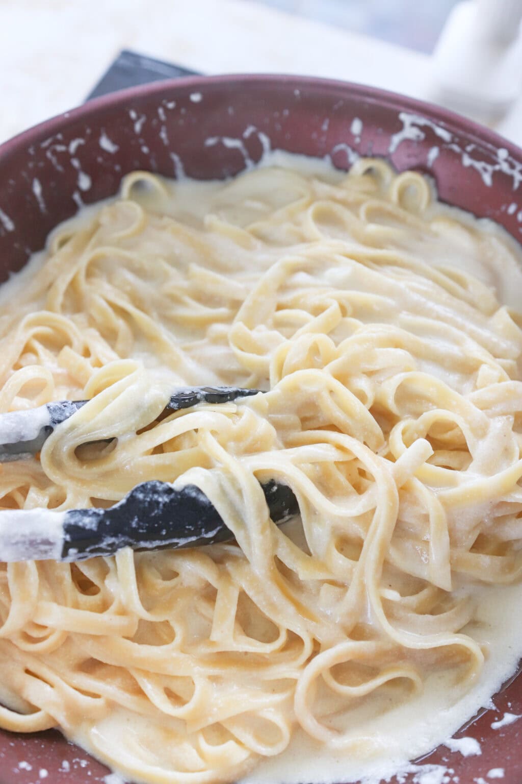 Fettuccine pasta being tossed in a rich and creamy cottage cheese Alfredo sauce using kitchen tongs in a large pan.