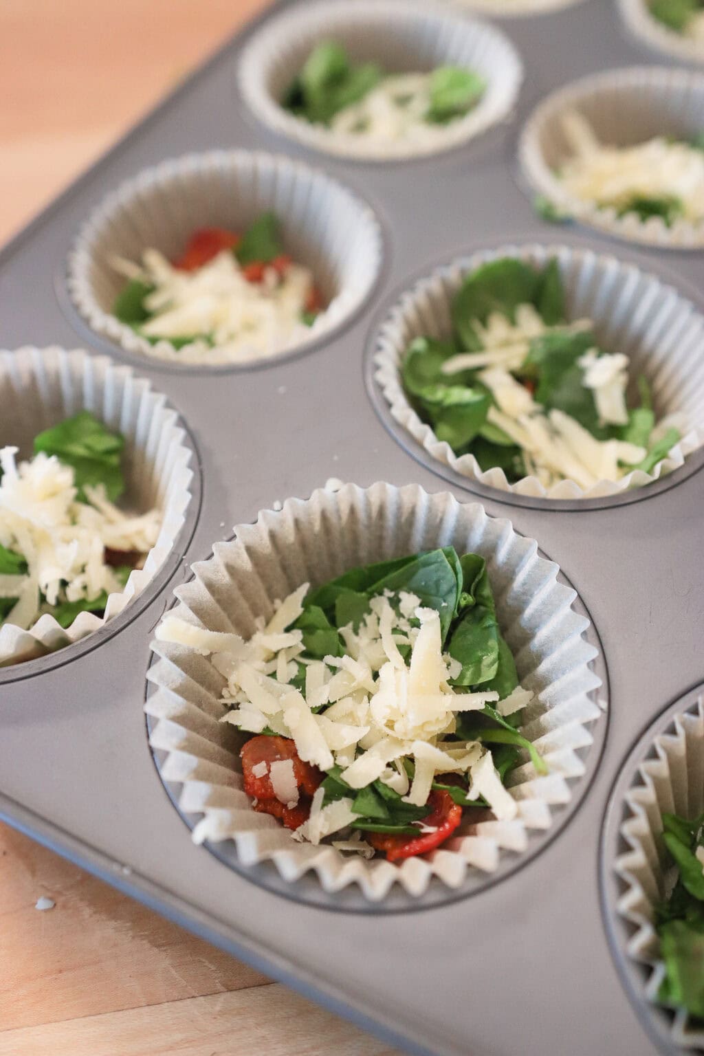 Close-up of muffin liners filled with fresh spinach, shredded cheese, and ingredients for egg bites, arranged in a baking tray.