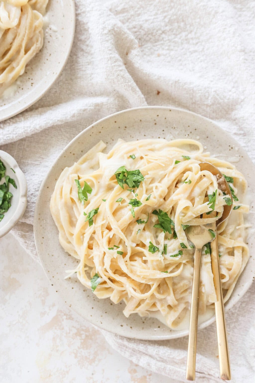 Two plates of cottage cheese Alfredo pasta garnished with parsley, served with golden utensils on a cozy textured cloth.