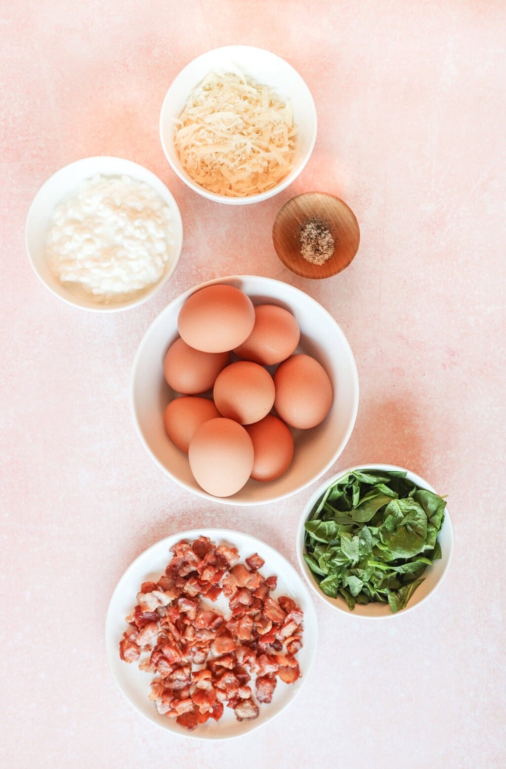 Flat lay of ingredients, including eggs, cheese, bacon bits, spinach, and cottage cheese, placed in white bowls on a pink surface.