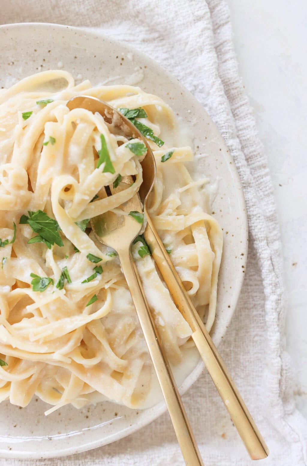 Close-up of a plate of fettuccine tossed in creamy cottage cheese Alfredo sauce, garnished with fresh parsley, with golden utensils resting on the side.