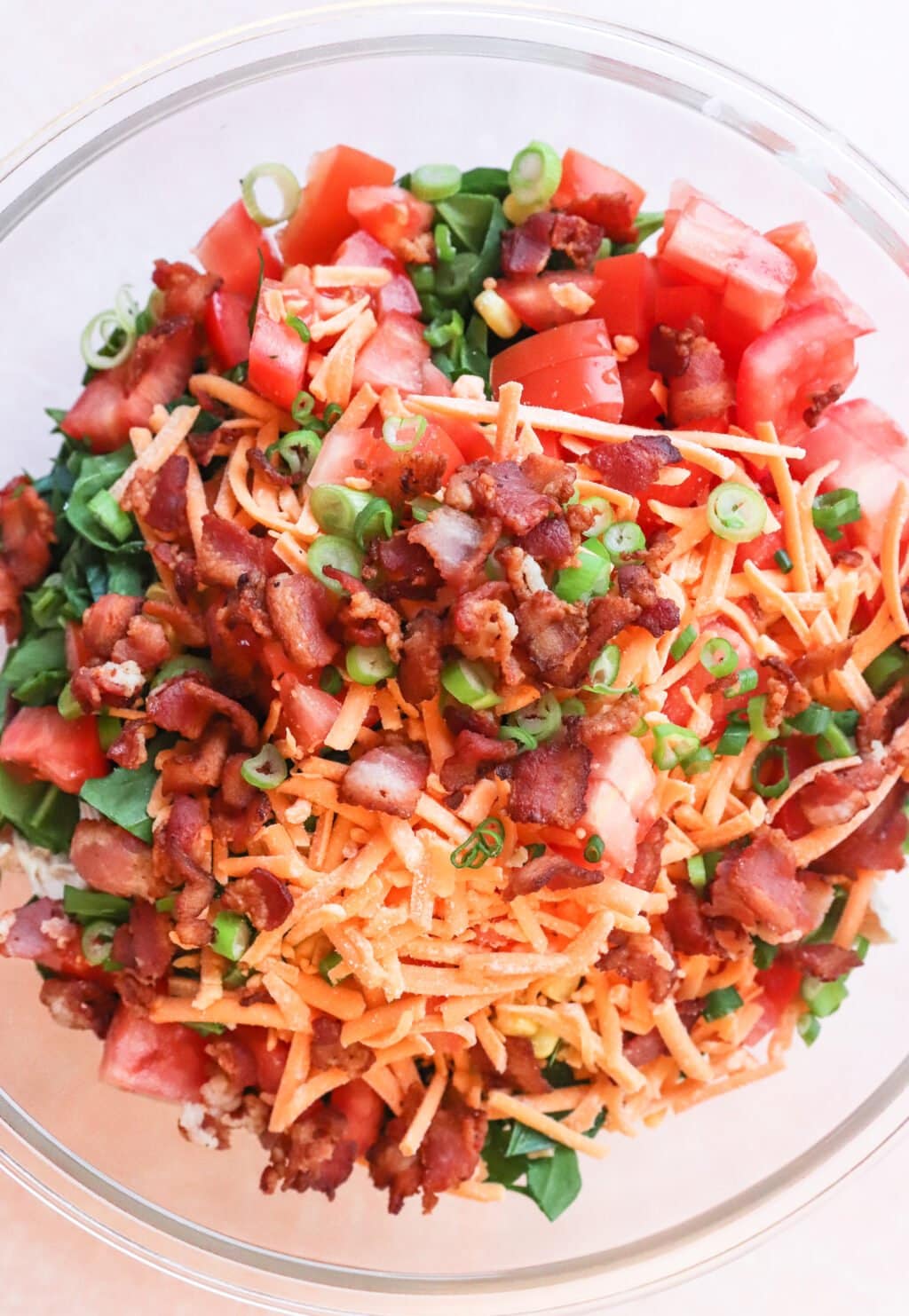 A glass bowl filled with a salad made of diced tomatoes, shredded cheddar cheese, chopped bacon, and sliced green onions on a bed of leafy greens.