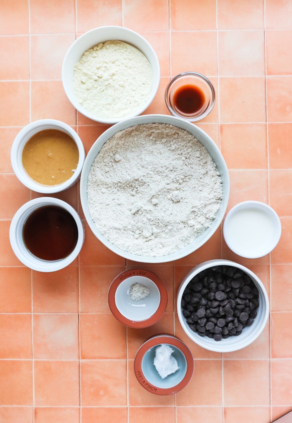 Bowls on a tiled surface filled with various baking ingredients: flour, chocolate chips, peanut butter, vanilla extract, milk, protein powder, a coconut oil portion, and a brown powder.