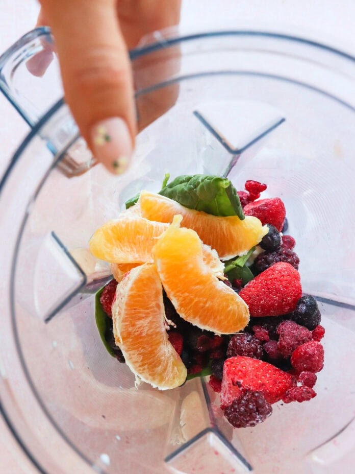 A hand holds a blender jar containing orange slices, frozen mixed berries, and spinach leaves, ready for blending.