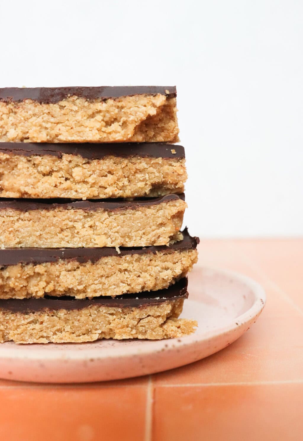 A stack of four chocolate-topped oat bars on a speckled ceramic plate. The bars have a thick oat base with a smooth, dark chocolate layer on top. Theyre sitting on a terracotta-colored surface against a white background.