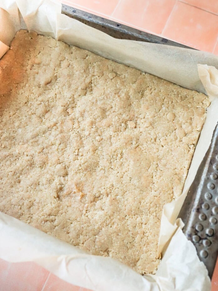 A rectangular baking pan lined with parchment paper holds an unbaked crumbly dough, evenly pressed into the bottom. The pan rests on a tiled surface.