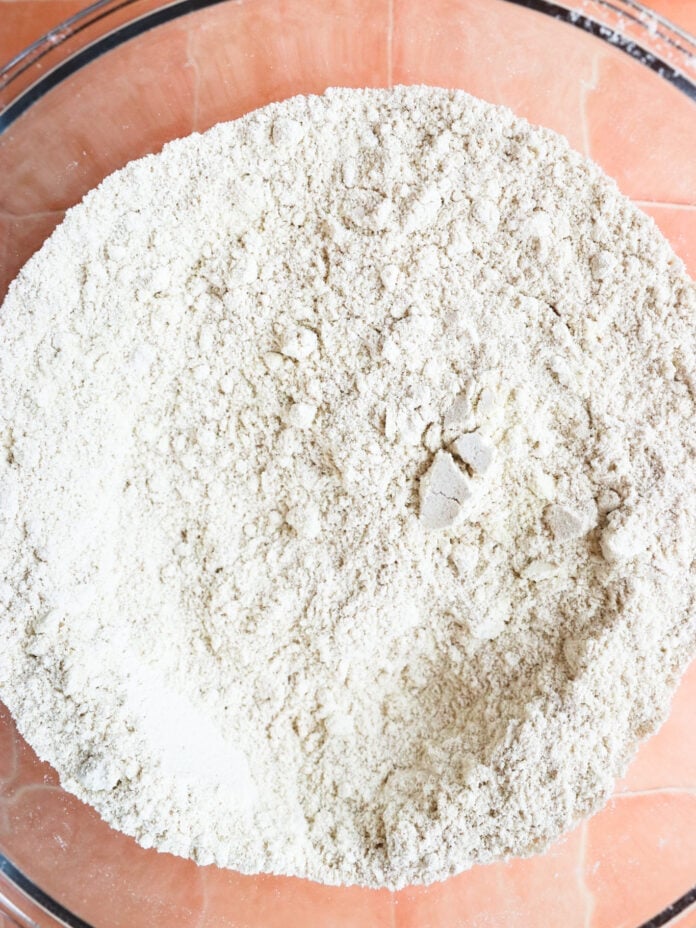 A close-up of a clear mixing bowl filled with pale, fine flour mixture on a terracotta-colored tiled surface. The flour is slightly unevenly distributed, forming small clumps and ridges within the bowl.