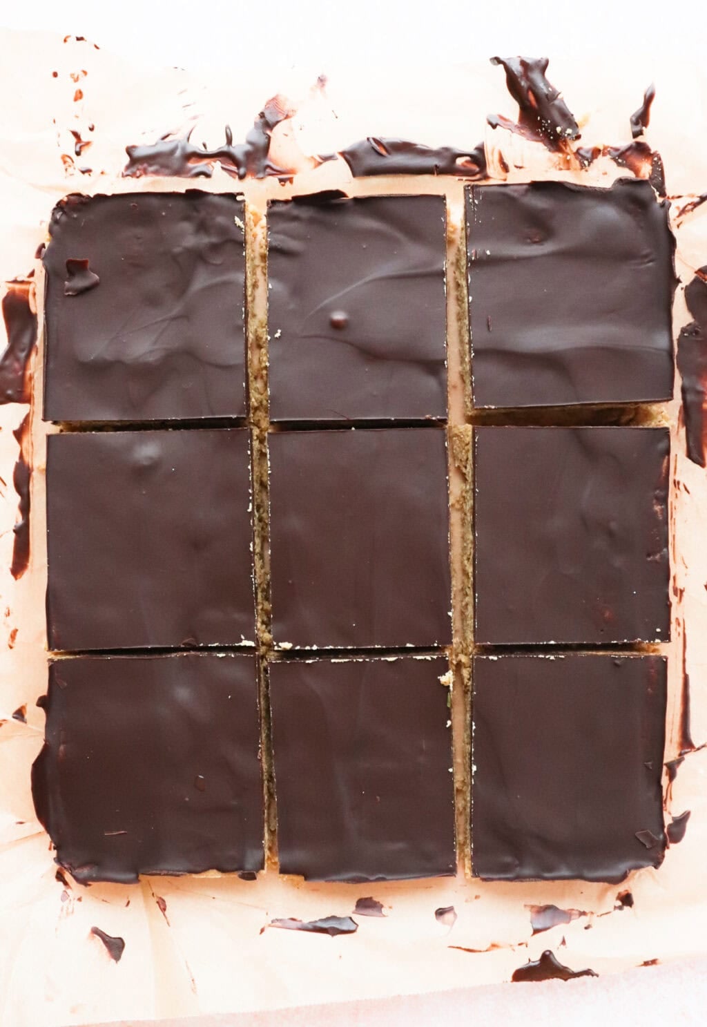 Nine square chocolate-topped dessert bars arranged in a grid on parchment paper. The bars have smooth, dark chocolate layers on top, and the edges are slightly uneven with a few chocolate smears on the paper.