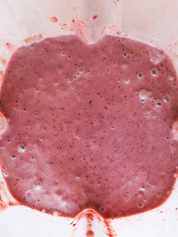 Close-up of a blender with a frothy pink smoothie inside. The smoothie has small bubbles on the surface and appears creamy. The interior of the blender has some streaks of smoothie along the sides.