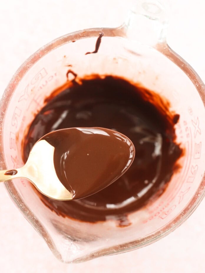 A glass measuring cup filled with melted chocolate, ideal for drizzling over homemade protein bars, features a gold spoon holding some of the rich chocolate above. The softly blurred background adds to the delicious anticipation.