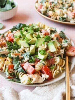A plate of pasta salad with rotini, diced tomatoes, avocado, chicken pieces, and chopped green onions, garnished with fresh greens. A fork rests beside the plate on a cloth, with another plate and a bowl of greens in the background.