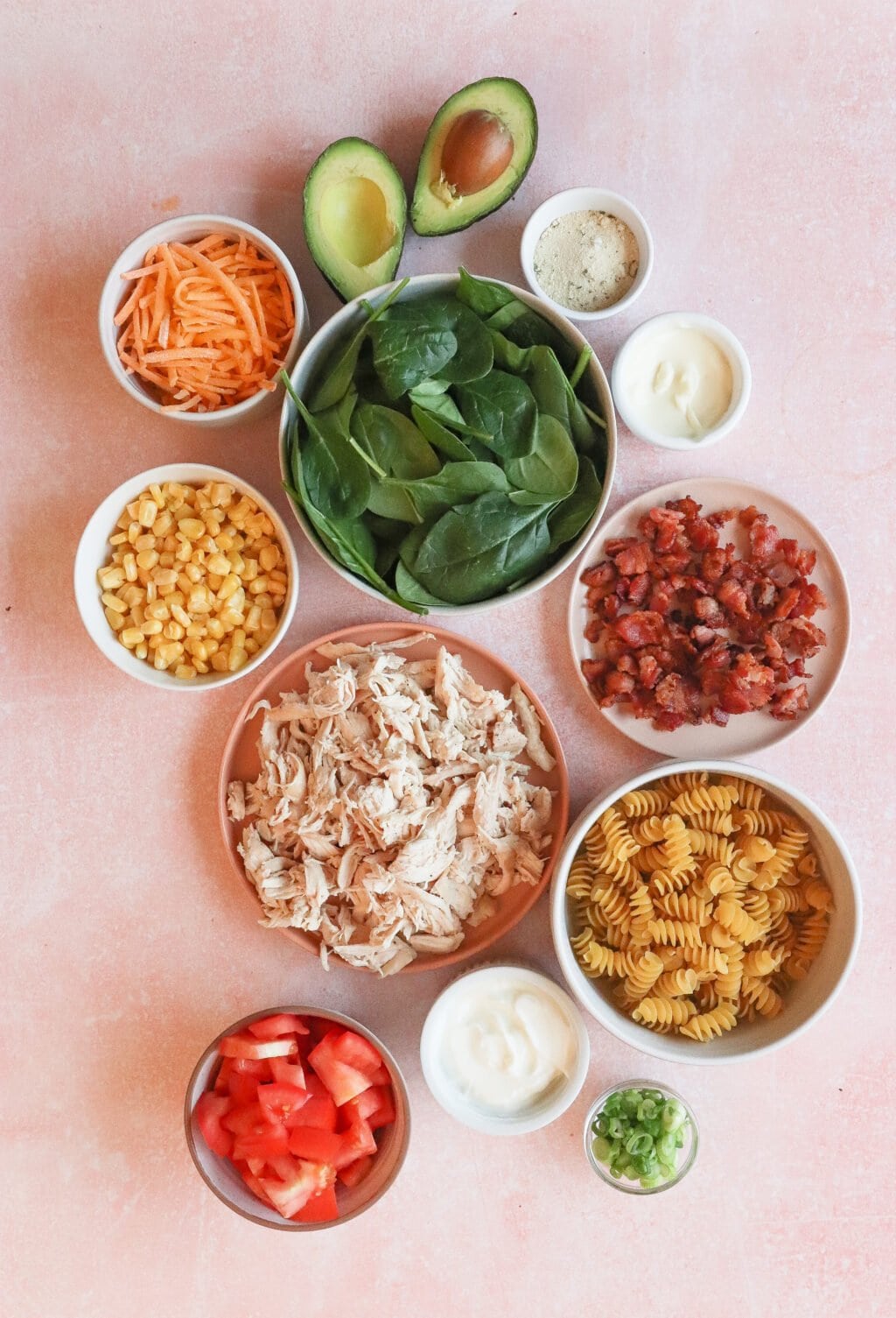 Ingredients arranged in a circle on a pink surface: spinach, avocado halves, sliced carrots, corn, shredded chicken, bacon bits, rotini pasta, diced tomatoes, green onions, and two small bowls of creamy dressing.