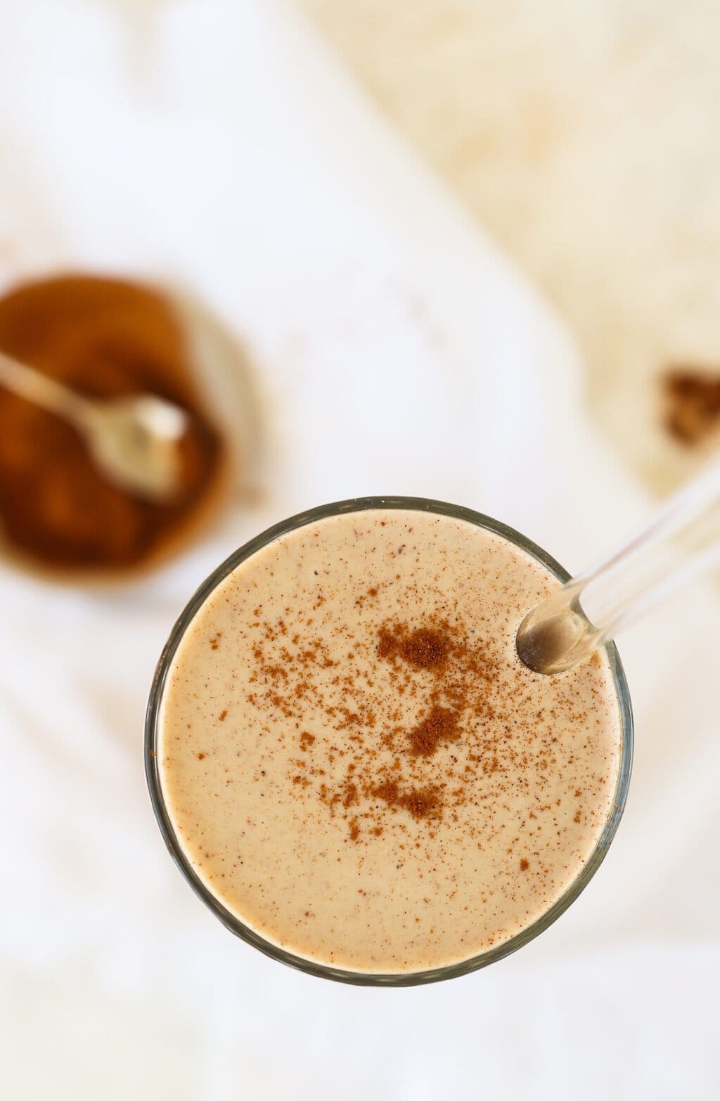 A close-up of a creamy beverage in a glass, topped with a sprinkle of cinnamon, with a glass straw inserted. A blurred bowl with a spoon is visible in the background.
